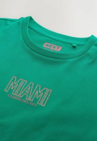 T-shirt vert col rond avec le texte « MIAMI CAMINO DEL REY » en lettres à motifs et une étiquette « NEXT RELAXED FIT » à l’intérieur du col.