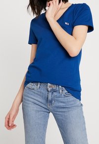 Royal blue cotton T-shirt with a v-neck, short sleeves, and a small logo patch on the left chest; paired with light blue jeans.