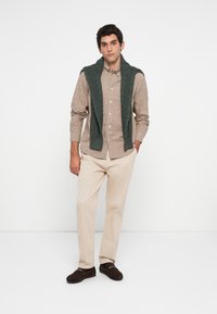 Man standing against white background wearing beige checked shirt, cream trousers, dark loafers, with dark green sweater draped over shoulders.