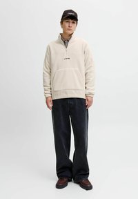 Young man wearing a beige quarter-zip fleece, black baseball cap, baggy black jeans, and brown shoes, standing against a plain white background.