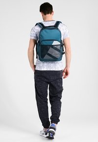 Ryggsäck i turkosa tyger med en strukturerad form, som har en framficka i nät och geometriska detaljer, bärs med mörka mönstrade byxor.