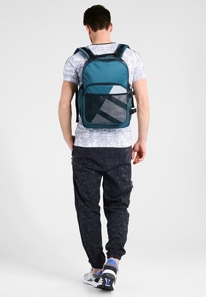 Batoh v tyrkysové látce se strukturovaným tvarem, s přední síťovanou kapsou a geometrickými akcenty, nošený s tmavými vzorovanými kalhotami.