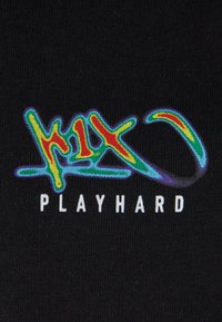 Sort stof med farverig, struktureret broderi. Grafikken inkluderer ordet "playhard" under et stiliseret logo med varierede farver.