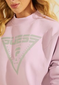 Felpa rosa chiaro con logo triangolare e la scritta "GUESS U.S.A. NINETEEN EIGHTY-ONE" in verde. Collo rotondo e morbida al tatto.