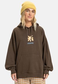 Brauner, übergroßer Hoodie mit einer Fronttasche, ausgestattet mit einem tan- und blauen Quiksilver-Logo. Kombiniert mit karierter Hose und einer gelben Mütze.