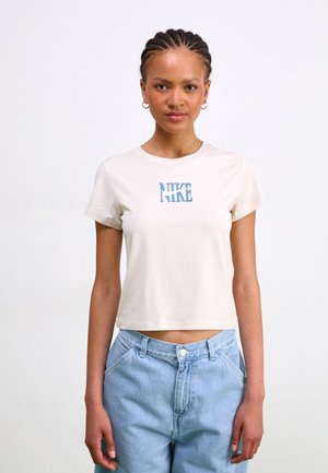 T-shirt crème à manches courtes avec un logo bleu "NIKE" centré sur la poitrine. Le tissu semble lisse ; le t-shirt a une coupe relaxée et cropped.