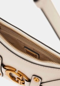 Beżowa skórzana torebka z metalowymi, złotymi elementami, wyposażona w zapięcie na suwak, kieszenie wewnętrzne oraz metkę z logo. Kontrastujące czarne akcenty na krawędziach.