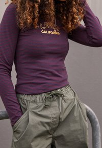 Lila Langarmshirt mit dünnen maroonfarbenen Streifen und einem goldenen Logo auf der Brust, kombiniert mit lockeren olivgrünen Shorts mit verstellbarem Bund.