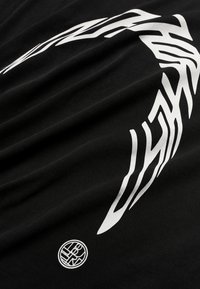 Μαύρο ύφασμα με λευκό στυλιζαρισμένο φυλετικό σχέδιο που κυκλώνει και ένα μικρό κυκλικό έμβλημα με αφηρημένα γράμματα κοντά στην άκρη.