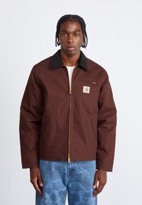 Brown jacket with a black collar, featuring a zippered front and chest pocket, crafted from durable material. Gold zipper hardware visible.