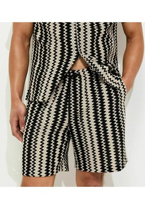 Uomo che indossa pantaloncini con motivo a zigzag bianco e nero e camicia a maniche corte abbinata, con una mano in tasca e in piedi davanti a uno sfondo bianco.