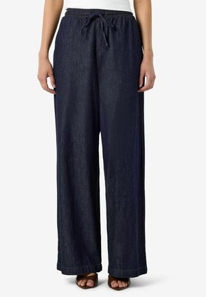 Femme portant un pantalon large en jean bleu foncé ample avec une taille élastiquée et un cordon, associé à des sandales marron ouvertes à l'avant.