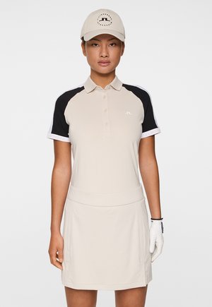 Biege golfpolo med svarta raglanärmar och vita accenter, med en knappslå, subtil logotyp på bröstet och matchande kjol. Golfhandske bärs på vänster hand.