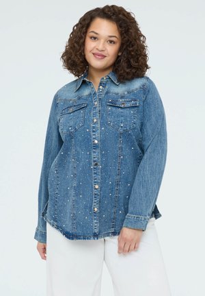 Žena s kudrnatými vlasy, oblečená v modré denimové košili s cvoky a bílých kalhotách, stojící před jednoduchým světlým pozadím.