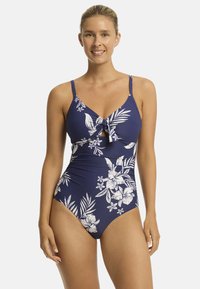 Vrouw die glimlacht, gekleed in een marineblauwe badpak met witte bloemmotieven. Staand tegen een egale witte achtergrond. Natuurlijk, helder licht.