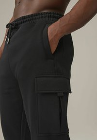 Pantalones de chándal negros hechos de un tejido suave, con una cinturilla elástica y cordón, bolsillos laterales y un gran bolsillo tipo cargador en el lateral.
