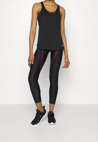 Svart idrottstopp i kombination med svarta leggings med lila sidostråkar. Modell som bär svarta sneakers. Slät tygtextur.