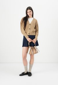 Beige strikkegenser med runde knapper, hvit topp, marinblå skjørt, beige ankelstrømper, svarte loafers og en flerfarget håndveske med kjedehåndtak.