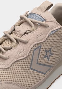 Κοντινή λήψη μπεζ αθλητικού παπουτσιού με επάνω μέρος από δίχτυ, επενδύσεις από σουέντ, μπεζ κορδόνια και ασημένιο λογότυπο αστέρα και σεβρόν στο πλάι.