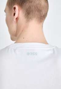 Fehér pamut póló, rövid ujjú, kerek nyakkivágással és egy kis, dombornyomott "BOSS" logóval a hátsó részen, a gallér közelében elhelyezve.
