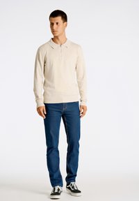 Beige pitkähihainen polo-paita, jossa on vetoketjullinen kaulus, yhdistettynä sinisiin farkkuihin. teksturoitu kangas, rento istuvuus ja mustat sneakersit valkoisilla pohjilla.