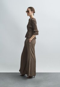 Brun plisseret bluse med lange ærmer og viddeærmer kombineret med brune bukser med vide ben. Modellen bærer oversized solbriller og står foran en neutral baggrund.