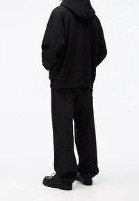 Personne portant un sweat à capuche noir ample, un jean large noir et des chaussures noires épaisses, debout de dos sur un fond blanc.