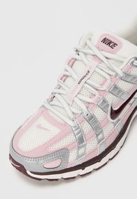 Nike tenisice s gornjim dijelom od bijele mrežice, ružičastim i srebrnim naglascima, podstavljenim jezikom i debelim potplatom. Imaju oznaku "B.Warm" na vezicama.