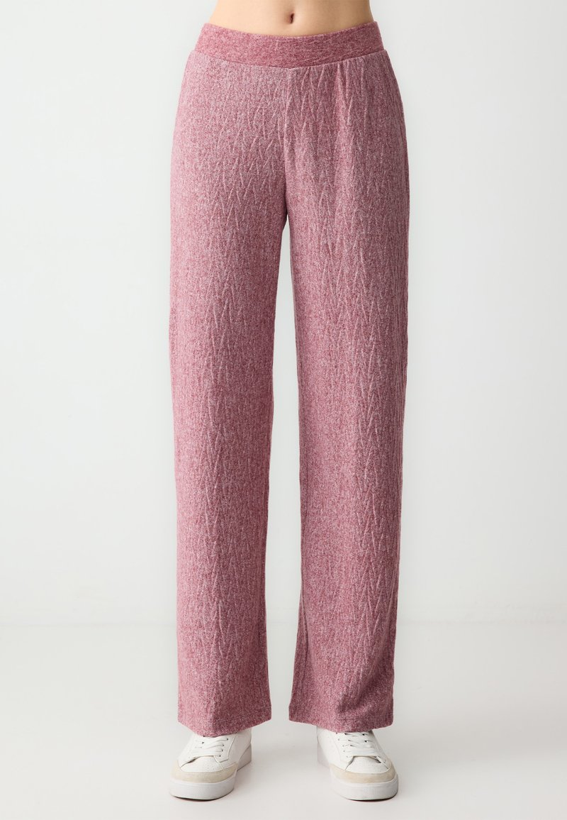 Różowe, fakturowane szerokie spodnie z geometrycznym wzorem i elastycznym paskiem w talii, w połączeniu z białymi sneakerami, na neutralnym tle.