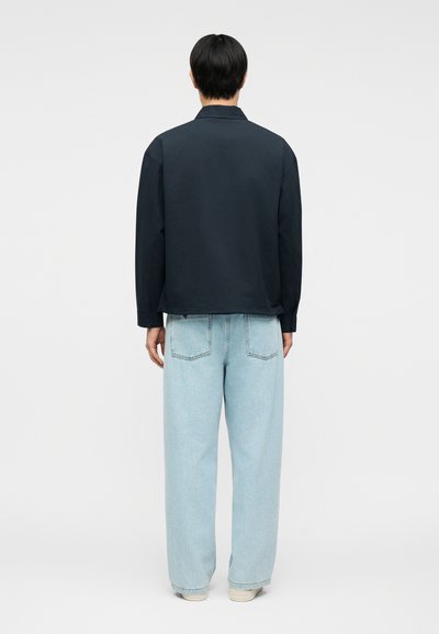 Άνδρας με την πλάτη προς τα εμπρός, φοράει σκουρόχρωμο πουκάμισο με μακριά μανίκια και ανοιχτό μπλε τζιν με φαρδιά πόδια, στέκεται μπροστά σε απλό φόντο.