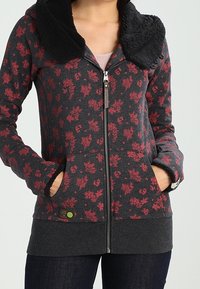 Dames zip-up hoodie in donkergrijs met een rood bloemenpatroon. Voorzien van een zwarte fuzzy kraag, twee voorzakken en geribde manchetten. Effen zoom.
