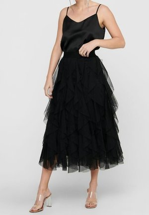 Woman wearing a black satin camisole with a layered black tulle midi skirt and clear high-heeled sandals on a light background.