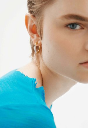 Pendiente de aro dorado con un acento blanco en un modelo con el pelo corto, que lleva una camisa azul rasgada. Enfoque en primer plano en la oreja y los detalles de la joyería.