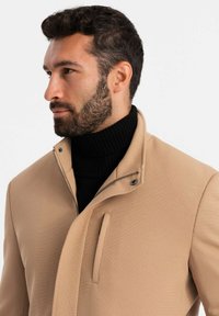 Cappotto beige testurizzato con colletto alto, con un dolcevita nero a costine sotto. Include una chiusura con zip e un dettaglio con tasca frontale.