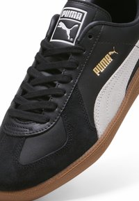 Czarne sneakersy PUMA z nubukową i skórzaną cholewką, białym paskiem po boku, brązową gumową podeszwą, płaskim sznurówkami i złotymi akcentami z logo.