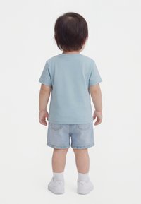 Småbørn står med ryggen til, iført en lyseblå kortærmet skjorte, denimshorts, hvide sokker og hvide sneakers.