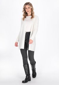 Cardigan lungo aperto in morbido tessuto avorio su una maglia crema, abbinato a jeans neri aderenti e stivali neri al ginocchio.