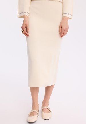 Kobieta w kremowej midi ołówkowej spódnicy i dopasowanym długim rękawem topie, w beżowych butach Mary Jane, stoi na białym tle.
