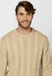 Pullover beige in maglia con lavorazione a trecce e scollatura tonda, caratterizzato da un motivo testurizzato e polsini a costine. Il tessuto appare morbido e spesso.