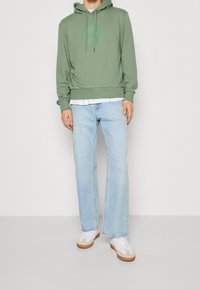 Person som bär ljusblå jeans, vita sneakers, grön huvtröja med subtilt tryck, och en vit skjorta under, står mot en plain bakgrund.