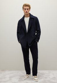 Marineblauw dubbelrijig jasje met zijzakken, gedragen over een lichte trui, gecombineerd met donkere corduroy broek en sneakers met beige accenten.