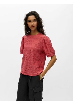 Femme aux cheveux bouclés et foncés porte un haut rouge à manches bouffantes ajourées et un pantalon cargo noir, debout avec une main dans la poche sur un fond uni.