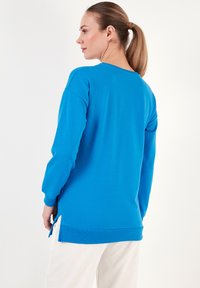 Sudadera azul de manga larga, con escote redondo y aberturas laterales en el dobladillo. Tejido suave con una textura lisa, llevada sobre pantalones de color claro.