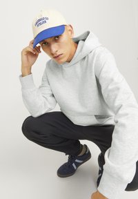 Jasnoszara bluza z kapturem z przednią kieszenią, zestawiona z czarnymi spodniami i ciemnoniebieskimi sneakersami z jasnoniebieskimi akcentami. Niebieska i kremowa czapka bejsbolowa.