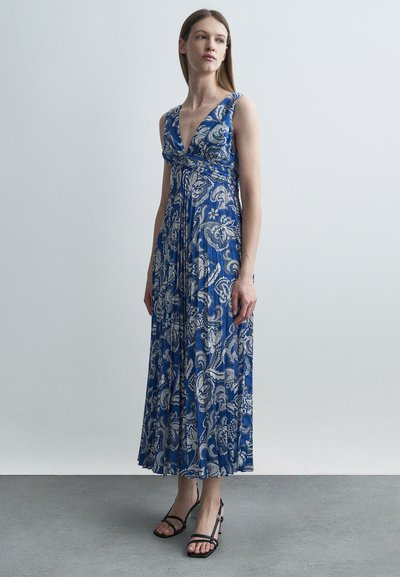 Femme debout portant une robe longue plissée sans manches, à fleurs bleues et blanches, et des sandales à talons noirs à lanières sur un sol gris.