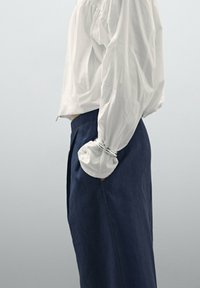 Blouse blanche à manches longues avec détail froncé aux épaules ; assortie à un pantalon taille haute bleu marine. Accent de manche en acier argenté.