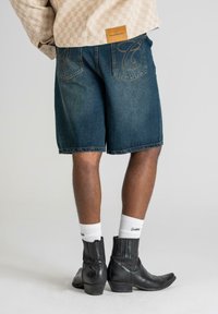 Shorts en denim bleu foncé avec une coupe décontractée, ornés d'un design cousu unique sur les poches arrière. Portés avec des bottines noires à talons pointus.