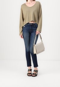 Suéter verde oliva recortado, jeans azul oscuro, sandalias negras con acentos dorados y un bolso gris claro. Diseño simple y estilo ajustado.