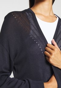 Mörk marinblå stickad cardigan med ett strukturerat mönster som har öppet arbete längs axlar och halsringning. Åtsittande ärmar.