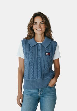 Donna sorridente con capelli ondulati che indossa un maglione polo senza maniche blu a trecce sopra una t-shirt bianca e jeans azzurri chiari, con la mano in tasca.
